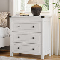 White farmhouse nightstand with 3 drawers and black metal knobs styled beside a bed with table lamp and books