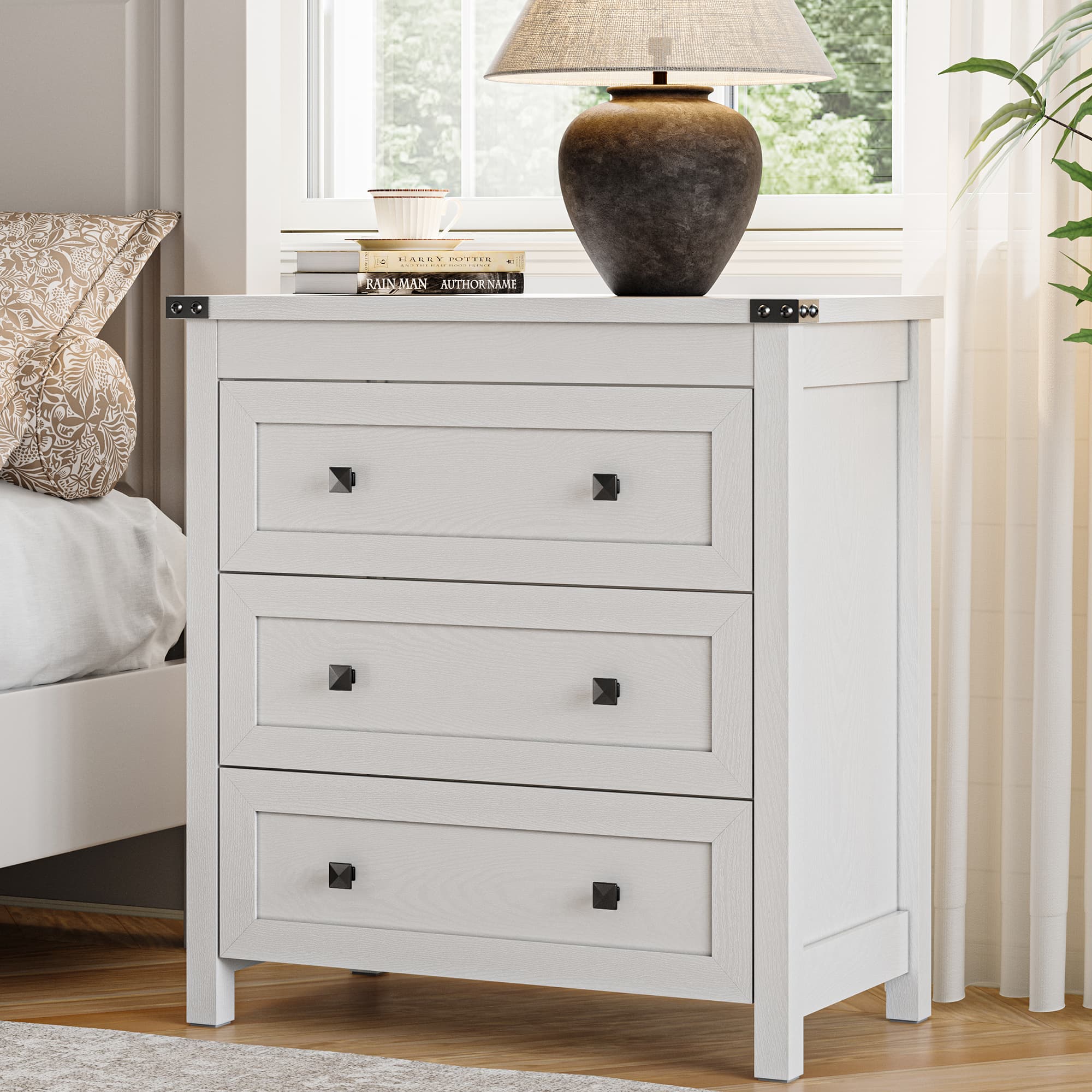 White farmhouse nightstand with 3 drawers and black metal knobs styled beside a bed with table lamp and books