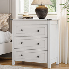 White farmhouse nightstand with 3 drawers and black metal knobs styled beside a bed with table lamp and books